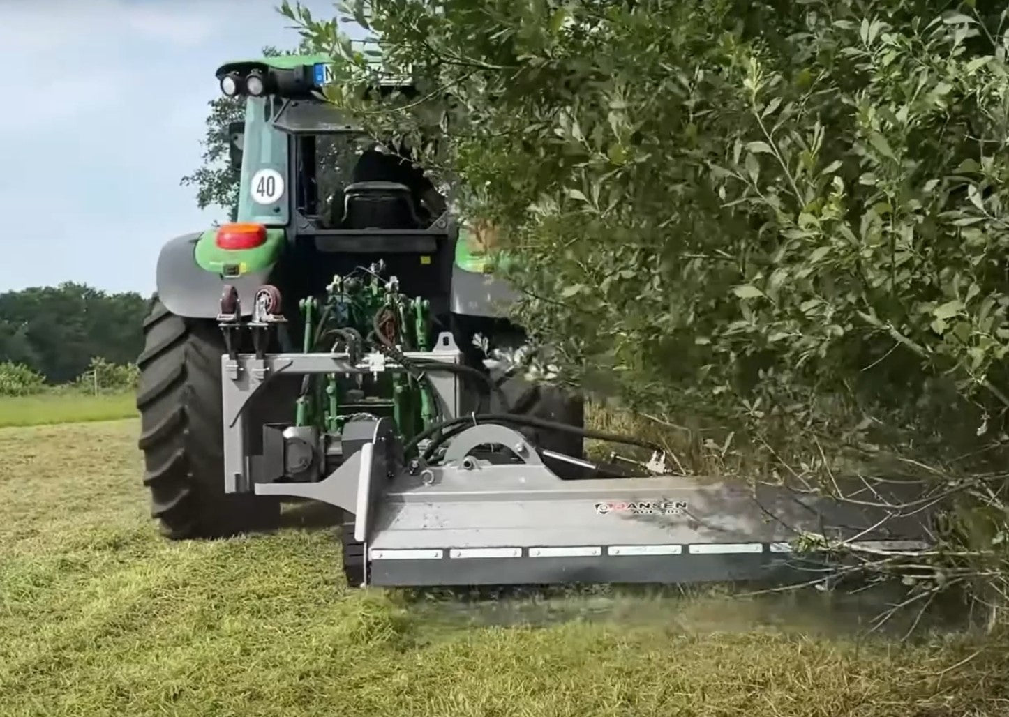 Rear view of a green tractor using a Jansen hydraulic verge mower to mulch a thick, overgrown hedge. Heavy-duty industrial machinery clearing dense vegetation on a grassy field with professional precision.